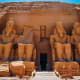 Three Egyptian pyramids in the Giza complex with a caravan of camels and riders crossing the desert sand under a blue sky wit